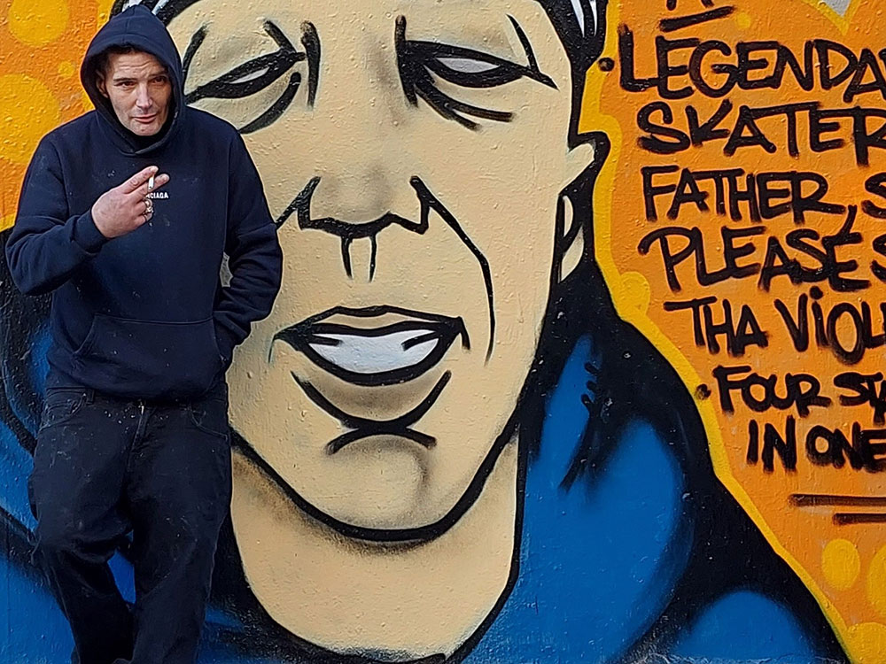 A man, Smokey D, stands in front of a mural of a young person in a blue hoodie and ball cap against a dark yellow background. Smokey Devil is wearing a blue hoodie, black jeans, and black and white skateboard shoes. He is holding a cigarette in his right hand.