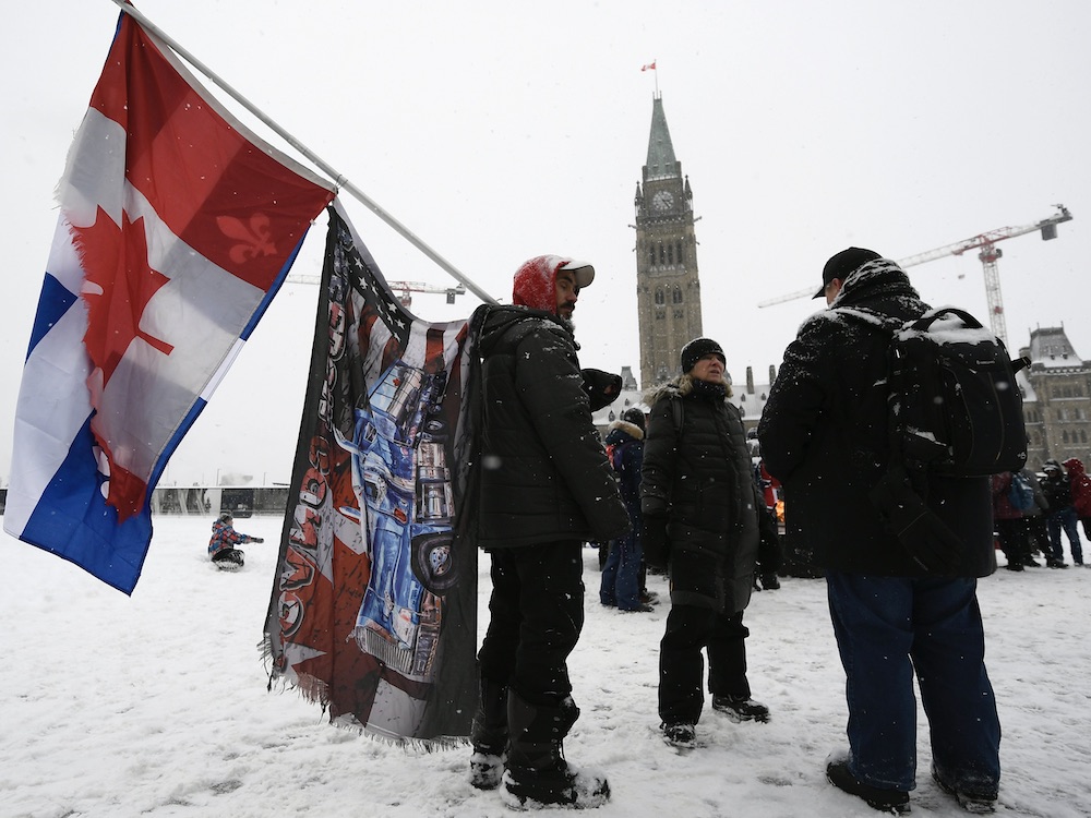 Three men in dark winter jackets stand in front of the Parliament buildings in Ottawa. One man in a ball cap and red hoodie is holding a set of flags. There is snow on the ground and on one of the men’s jackets. The sky is white.