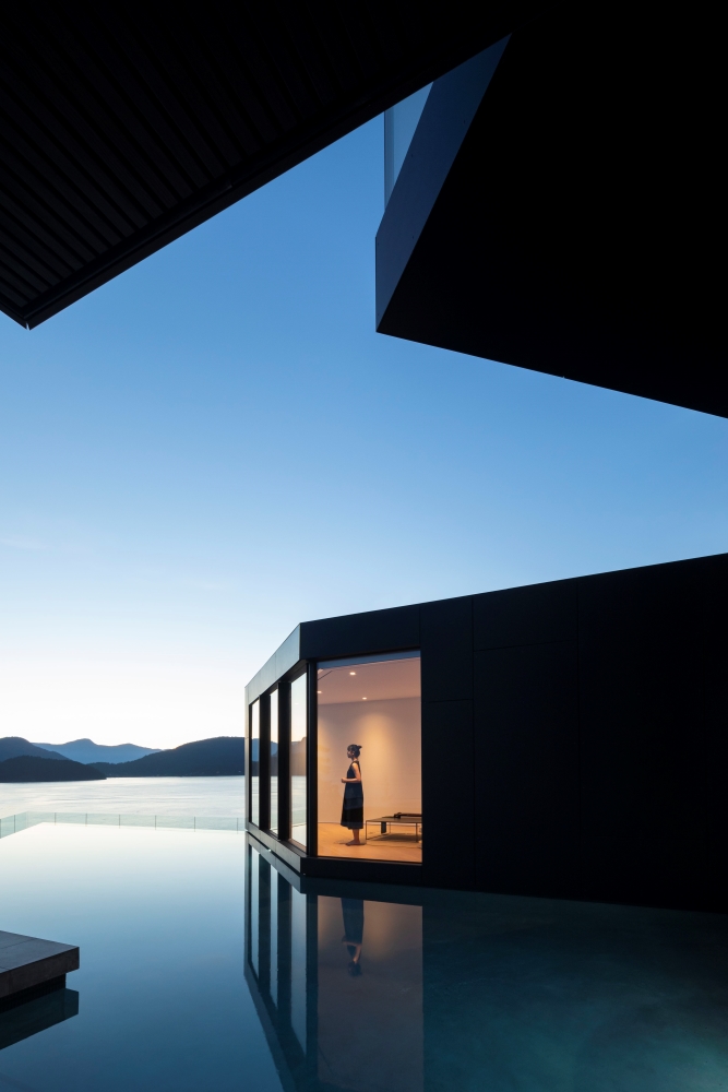 A vertical photograph depicts a large home on the water at dusk. The sky is blue and there are mountains in the background. A row of windows abuts the water, and a young woman in a black dress is standing in the window, looking out at the water towards the left of the frame.