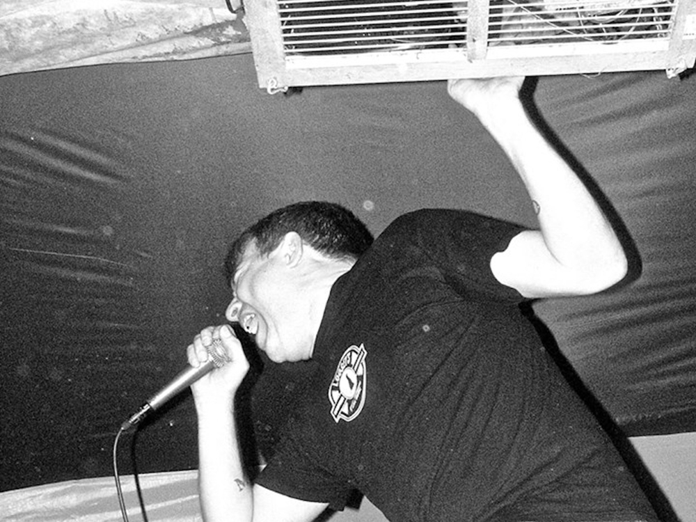 A black and white photo shows Jason Schreurs on stage, hunched over, singing intensely into a microphone. His other hand clutches a piece of HVAC equipment above his head. 