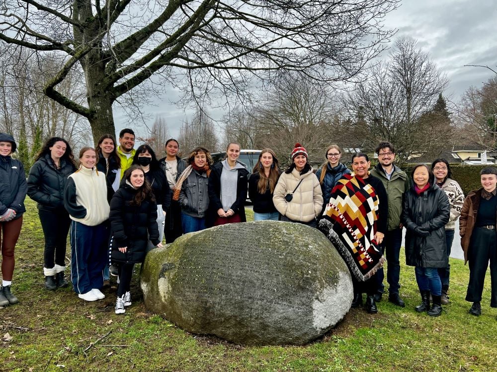 A group of young adults in winter clothing stands around a boulder on grass under a deciduous tree in a residential area. They are looking at the camera and smiling.