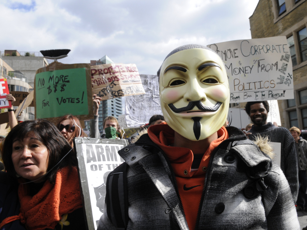 A person wearing a Guy Fawkes mask looks at the camera amid a protest on corporate investment in politics. Signs read 'Remove corporate money from politics” and “No more $$$ for votes!'