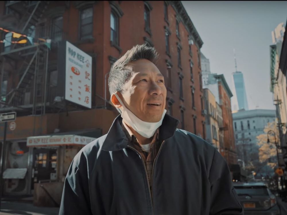 A middle-aged Chinese man partially turns his head towards the horizon to the right of the frame, smiling slightly. A white medical mask hangs across his neck. He is wearing a navy jacket and standing on a street corner in New York City in front of a brownstone apartment building on a sunny fall day.