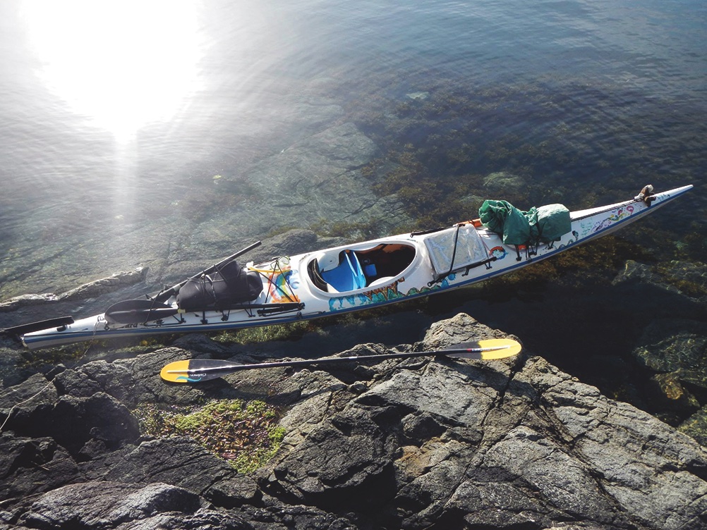A white kayak loaded with gear and decorated with small colourful paintings sits on a craggy Pacific shoreline on a sunny day. A corona flare to the upper left quadrant of the frame is reflecting off the clear, bright water to reveal rocks and plant life under the surface.