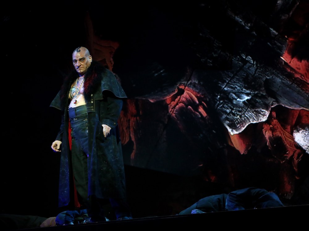 A bald man with a curly lock of black hair stands to the left of the frame. He is wearing a long, fur-lined black jacket over his bare tattooed chest, over which hangs a gold chain and dark red gem. The stage background against which he stands is black with thick painted red and white accents.