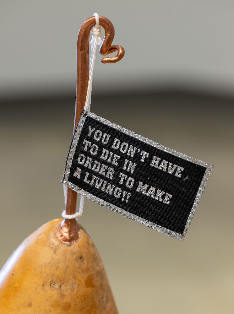 The vertical bronzed stem at the top of a pear-shaped golden gord holds a black flag with silver text in capital letters that reads, 'You don’t have to die in order to make a living!!'