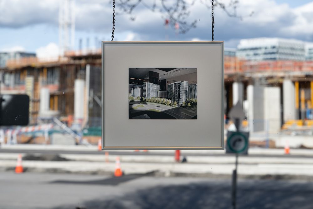 A small architectural photograph of a new condo development hangs by a silver chain in a silver frame against a window that looks across the street to a development in process. 