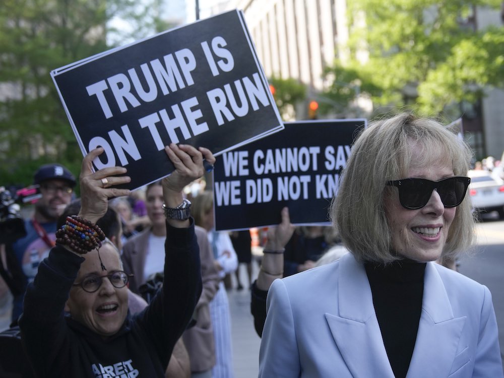 A woman with short blonde hair, sunglasses and a light grey blazer over a black turtleneck walks down a New York City sidewalk, smiling. Behind her, demonstrators hold black placards with white sans-serf typeface that read “Trump is on the run” and “We cannot say we did not know.”