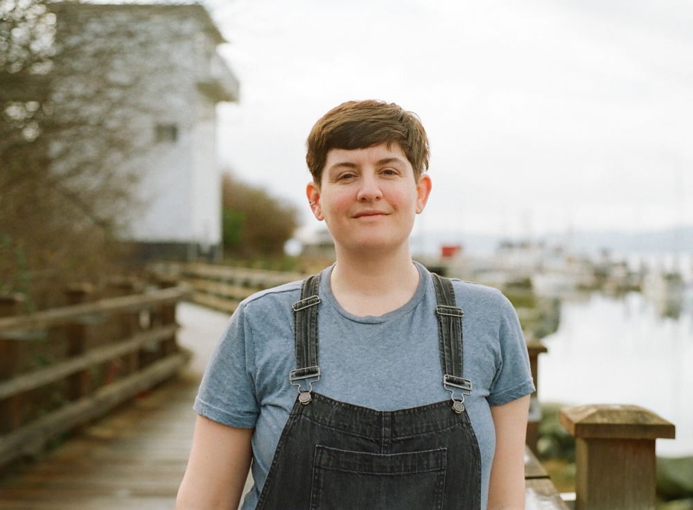 In focus, a non-binary person wearing black denim overalls stands on a bridge. Out of focus, a wooden bridge leading to a gravel path, from which rises a tall, skinny white building.