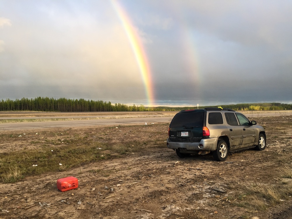 A grey sports utility vehicle sits on the side of the road. Behind it, a red jerry can of gas sits empty on its side. On the horizon against a grey sky, two rainbows are visible.