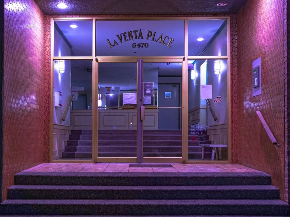 The entryway of an old apartment building, built sometime between the 1960s and ’70s. The image has a purple tint. La Venta Place is written above the glass doorway in a sharp-looking font. There is something eerie about the scene. 