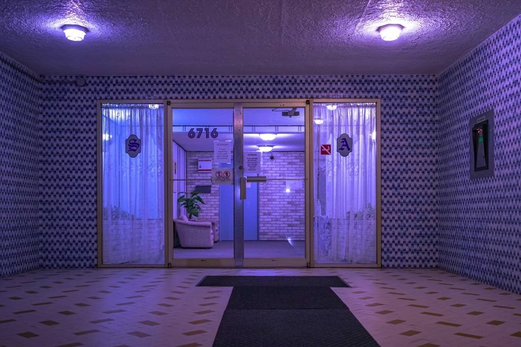 The entryway of an apartment building at night. There is a purple tint to the image. The lobby has brick walls and there is a satin couch beside a plant. The outside of the doorway is uncover and enveloped with glossy tile.