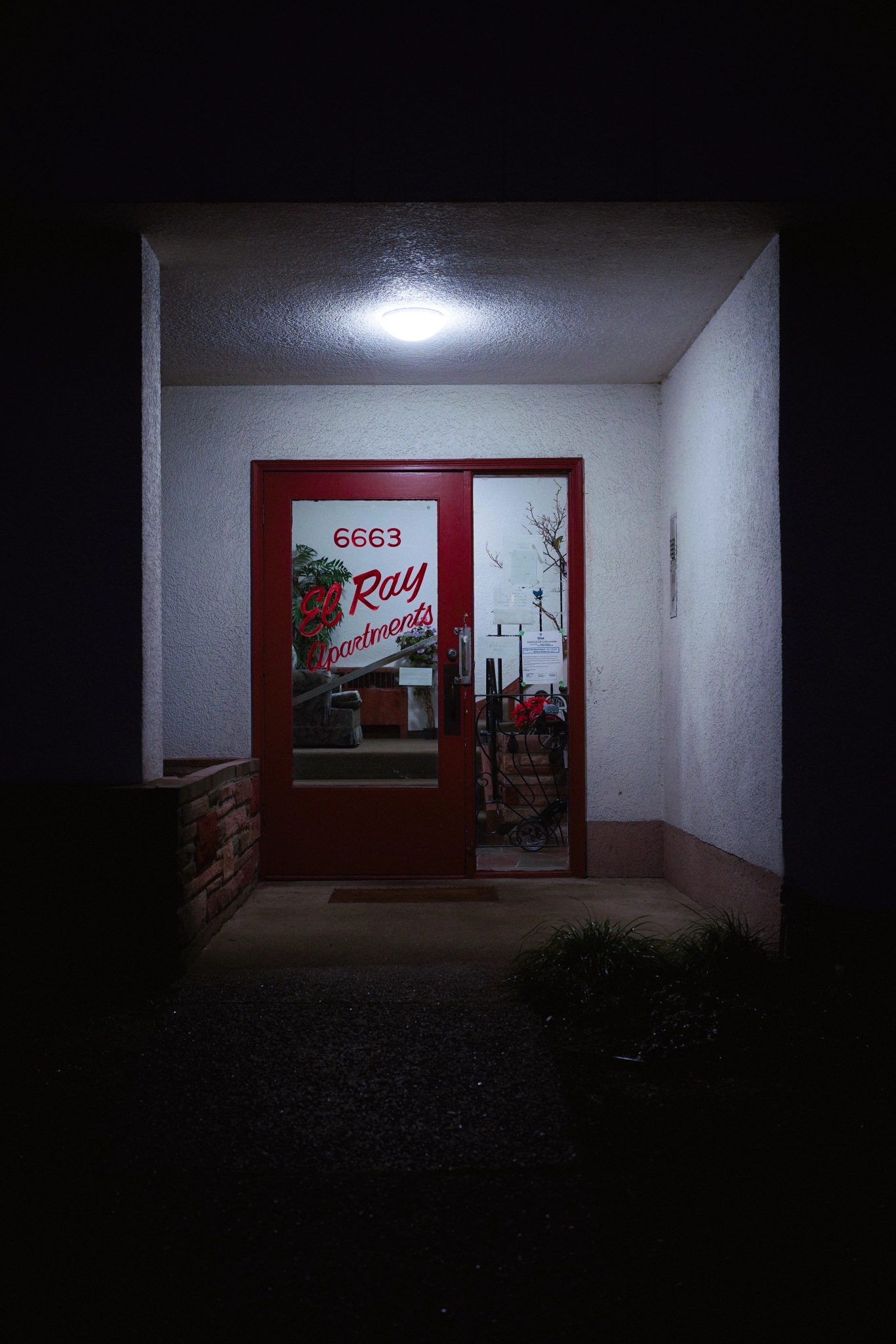 The entryway of an apartment building at night. The walls are stucco. On the door, the name “El Ray Apartments” is painted on in red in beautiful cursive.
