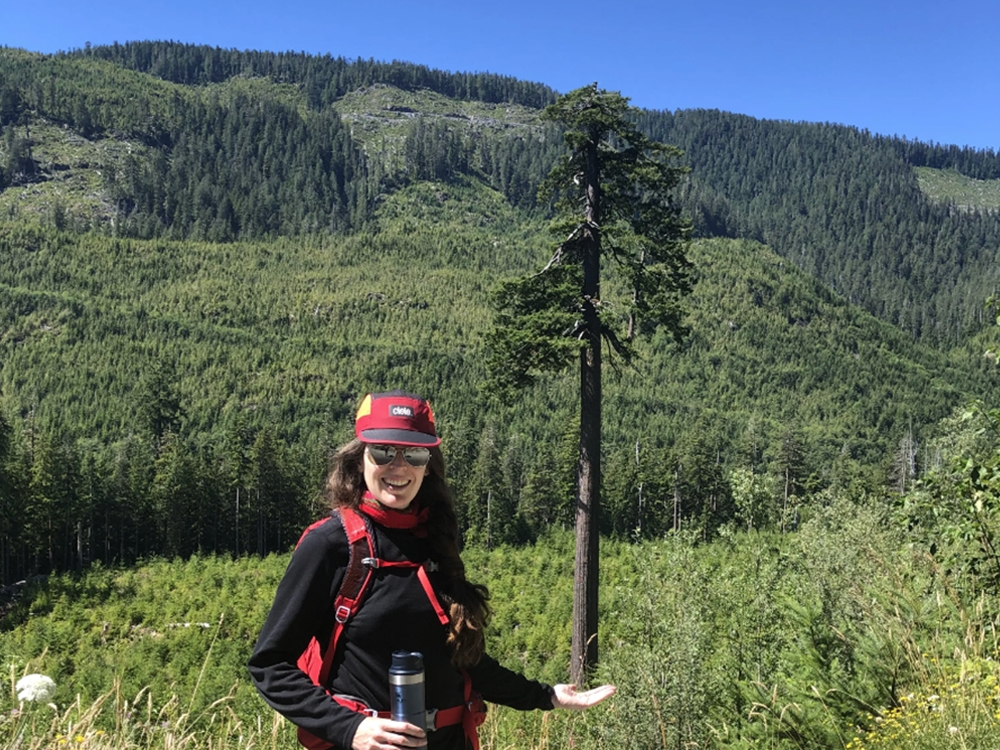 A woman wearing a hat and sunglasses extends her palm, so that it looks like a giant Douglas fir is sitting in the palm of her hand.
