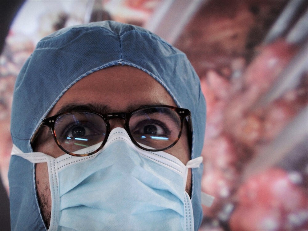 A man with glasses wears a blue medical mask and a blue surgical hood. In the background is a digital image of a medical procedure.