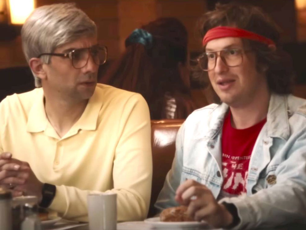 Two men sit next to each other in a diner booth. On the left is a young man with short grey hair, glasses and long-sleeved butter-yellow polo shirt. On the right is a man with longer brown hair, a red headband, glasses and a light blue denim jacket over a red graphic T-shirt. The man on the left is looking at the man on the right while he is speaking.