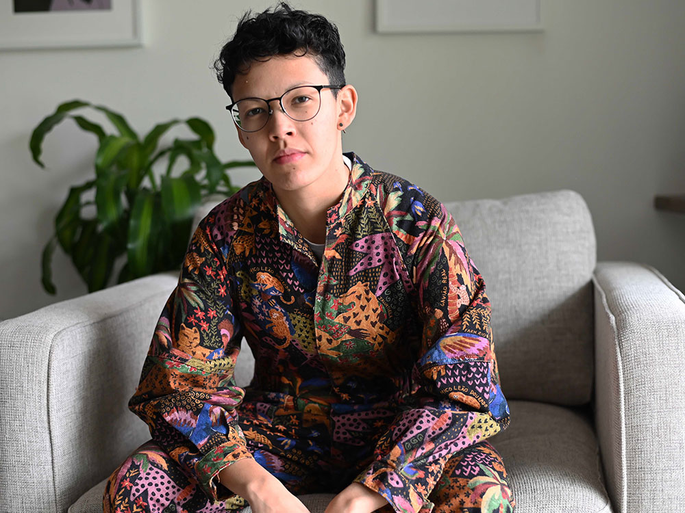 Emi Sasagawa sits on a light grey armchair. She has short dark curly hair and is wearing glasses and a colourful patterned jumpsuit. She is looking at the camera with her head tilted slightly.