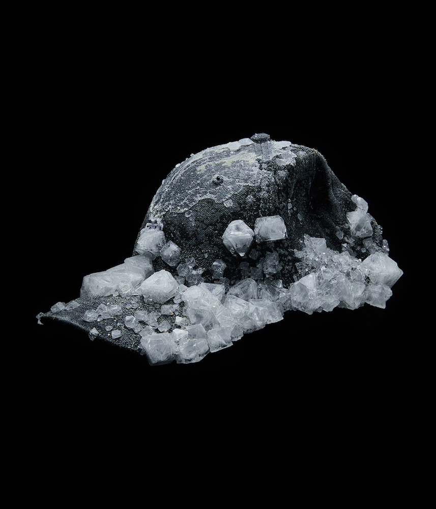 A black ball cap is encrusted in appliqued white plastic that resembles ice cubes. It is photographed against a black background.