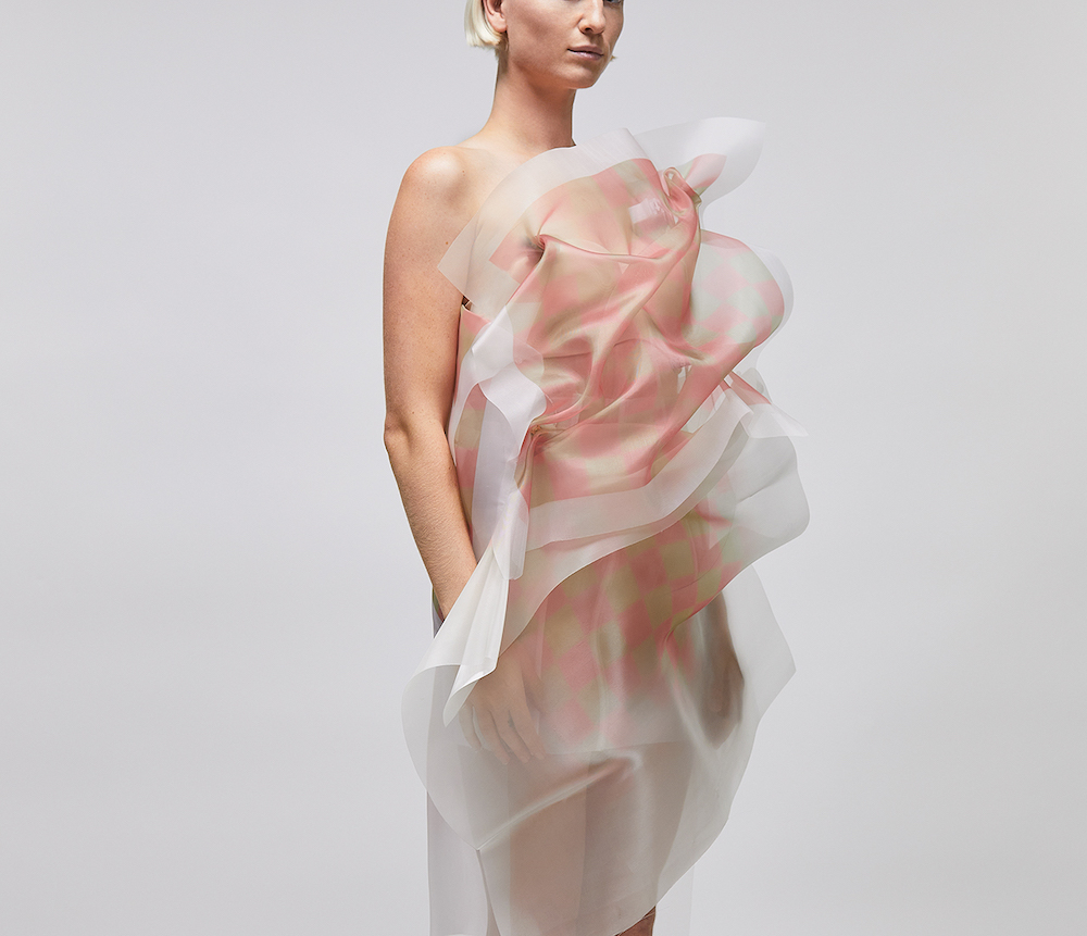 A woman with short blonde hair models an off-the-shoulder dress made of transparent, billowy fabric. We can’t see her eyes, and she stands against a light grey background.