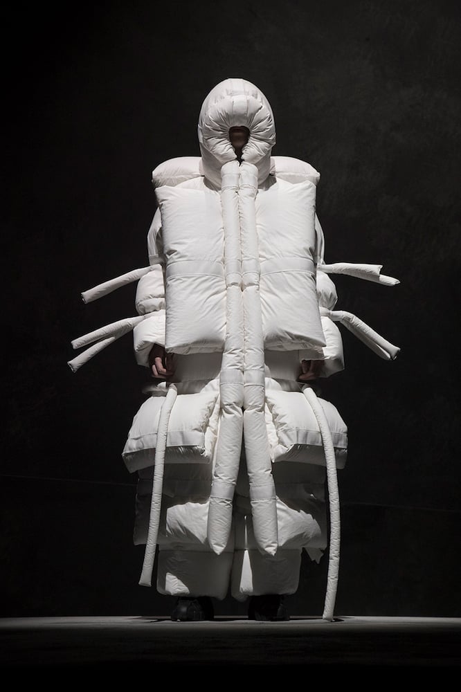 A model stands against a dark grey background in a full-body oversized puffy white jacket with white tubes extending from the elbows down the sides of the body. Their face is barely discernible inside the recesses of a deep hood.