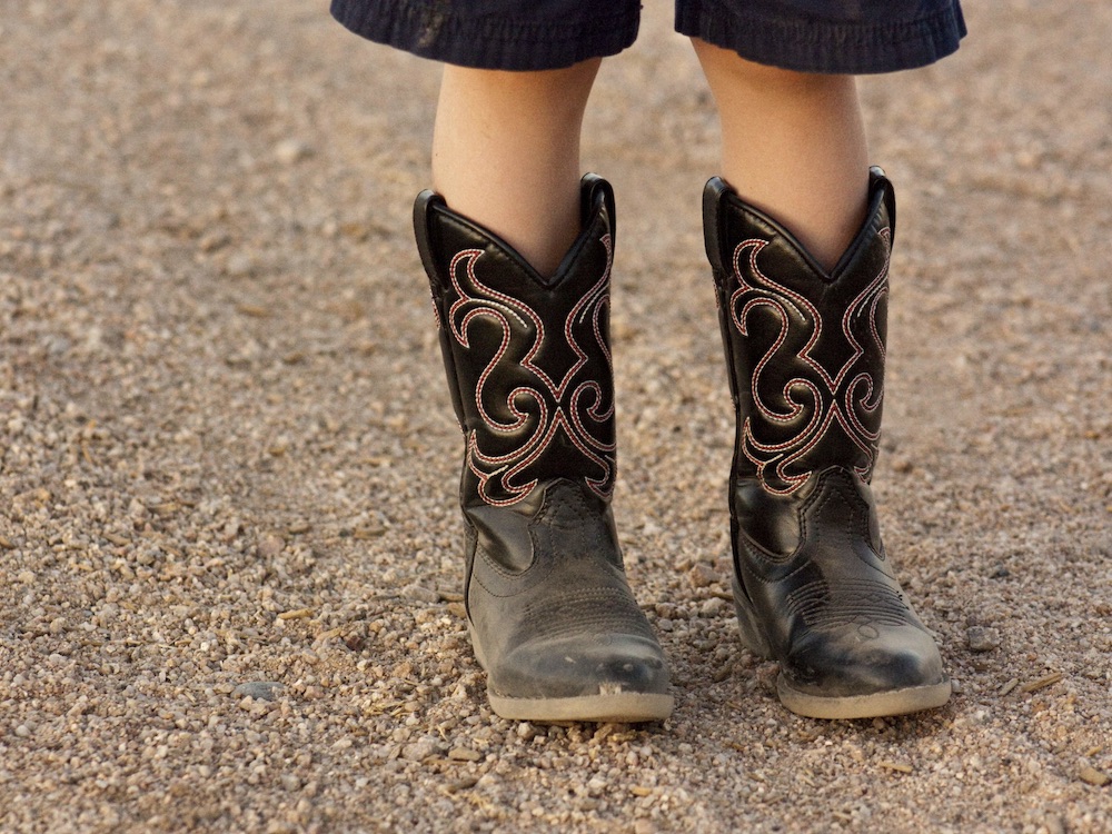 A child stands on a gravel road in a pair of black cowboy boots. They are wearing black shorts that stop at the knee. We can’t see the child above their knees.