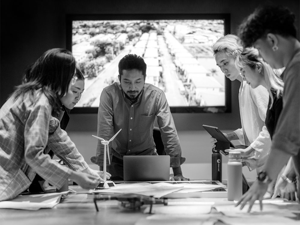 A black and white photo of a group of people gathering around a table with a screen behind them. A windpower mockup is on the table. They appear to be thinking critically.