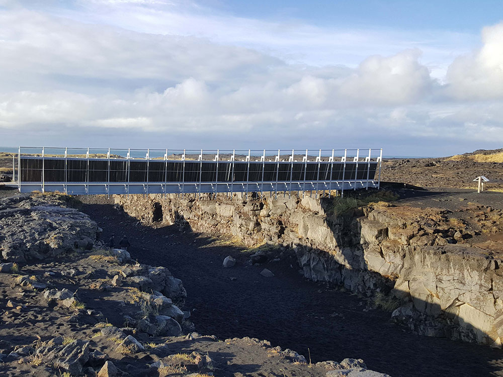 A steel pedestrian bridge spans across a rocky chasm in Iceland on a sunny day, where the sky is blue with large puffy clouds.