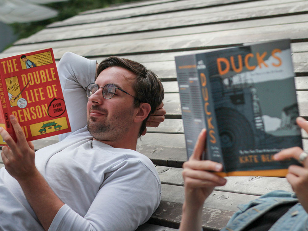 Two people recline, reading books: Kevin Chong’s ‘The Double Life of Benson Yu’, and Kate Beaton’s ‘Ducks.’