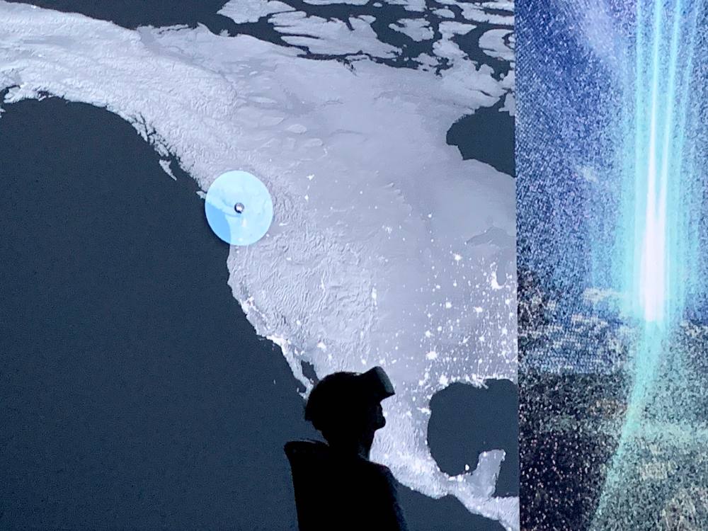 The silhouette of a person wearing a virtual reality headset is looking up towards a digital map of North America next to an artful digital rendition of fine particles in blue light.
