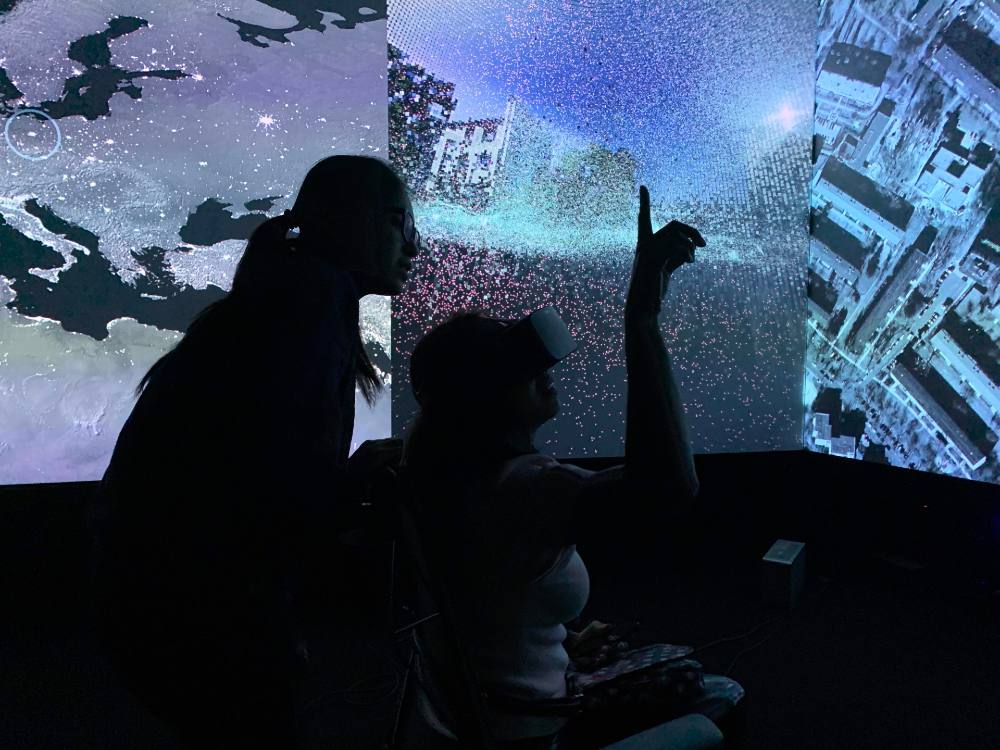A silhouetted person in glasses and a ponytail stands behind another person seated and wearing a virtual reality headset. The seated person is pointing upwards. Behind them is a wide, colourful digital installation featuring maps, a street-level photo of apartment buildings and an aerial photo of a city.