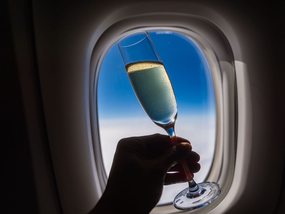 A silhouetted hand holds a champagne flute against an airplane window against a blue sky.