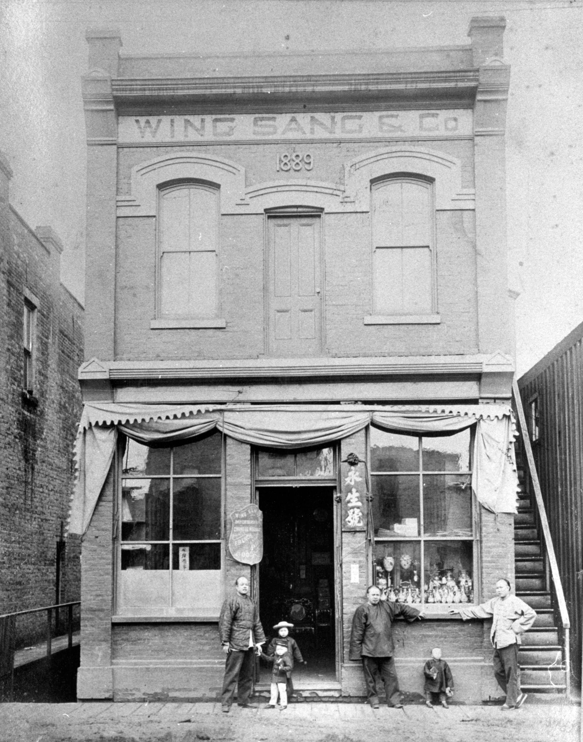 A man beside two children in the doorway of a two-storey Victorian commercial building. Two other men and a child stand in front of a packed window display. They are all ethnically Chinese. The photo is black and white.