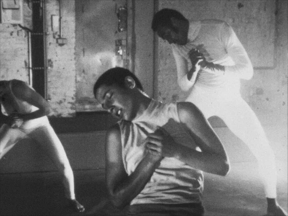 A black-and-white digital video still depicts a grainy image of three dancers in a studio. They are wearing light-coloured streetwear and leotards, bending towards the left of the frame.