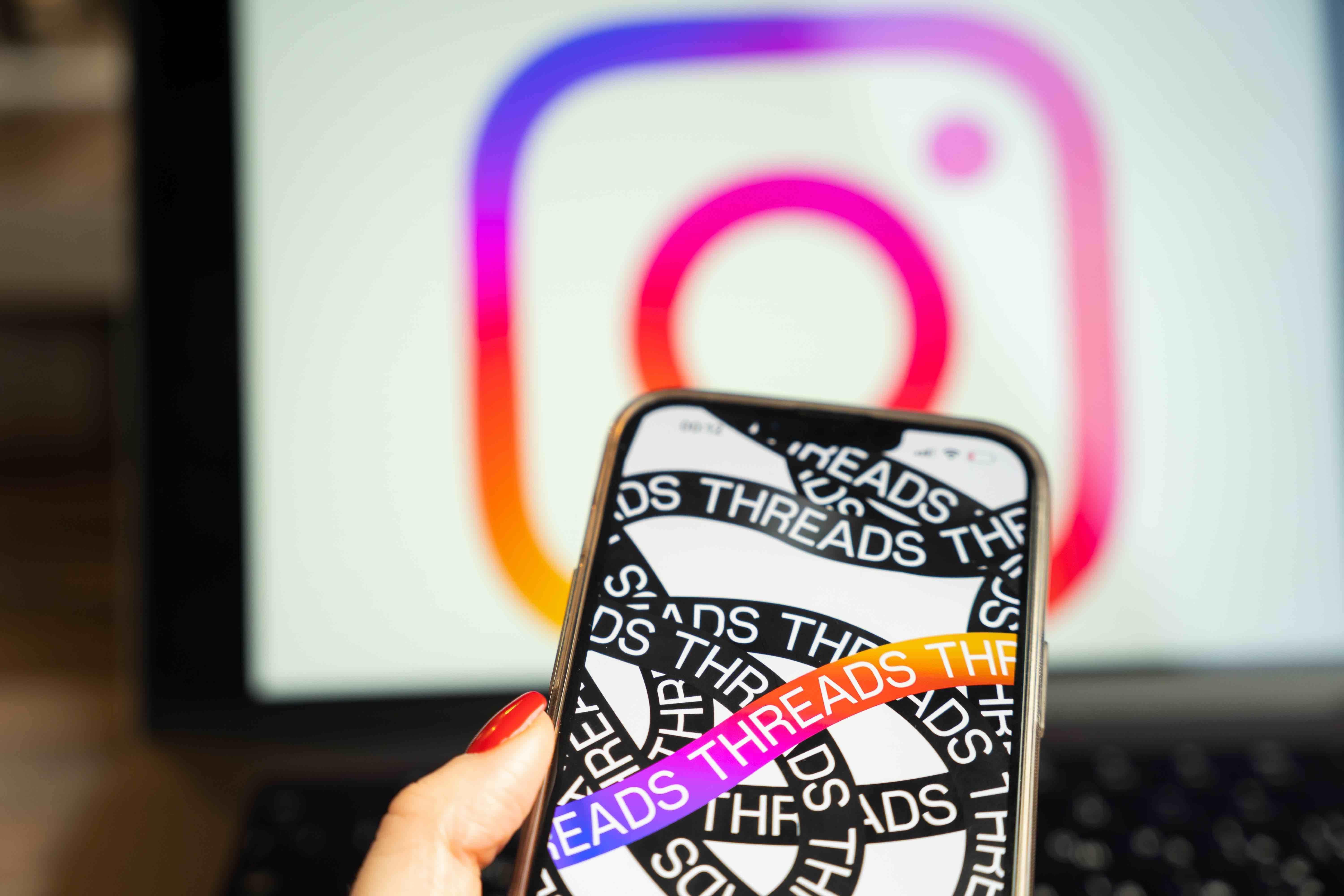 A manicured hand holds a smartphone featuring the Threads app. Behind the phone is a blurry image of the purple and orange Instagram logo. 
