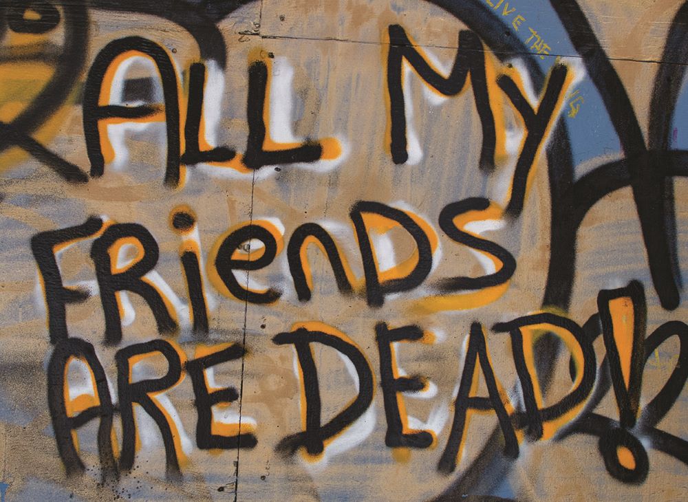 Graffiti on a wall says ‘All my friends are dead!’ The colours are vibrant, with black letters shaded with orange and white.