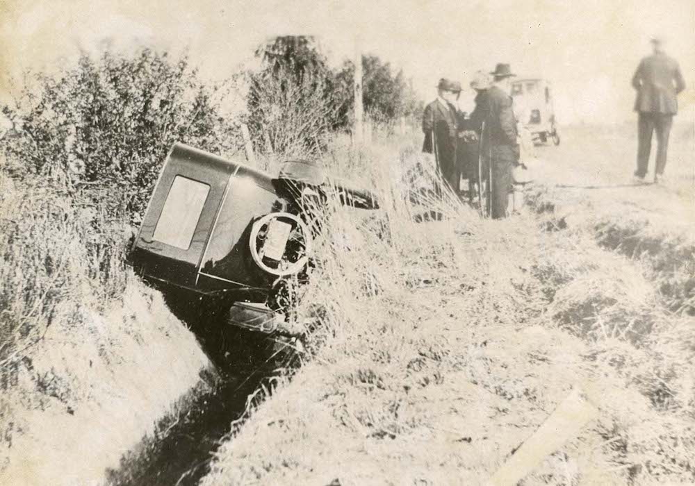 An old car in a ditch with formally dressed men by the side of the road.