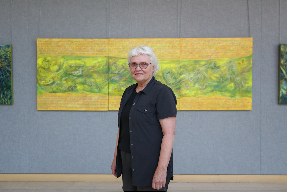 A woman with white hair stands in front of a large horizontal painting of textured ditch water, with cursive text above and below it.