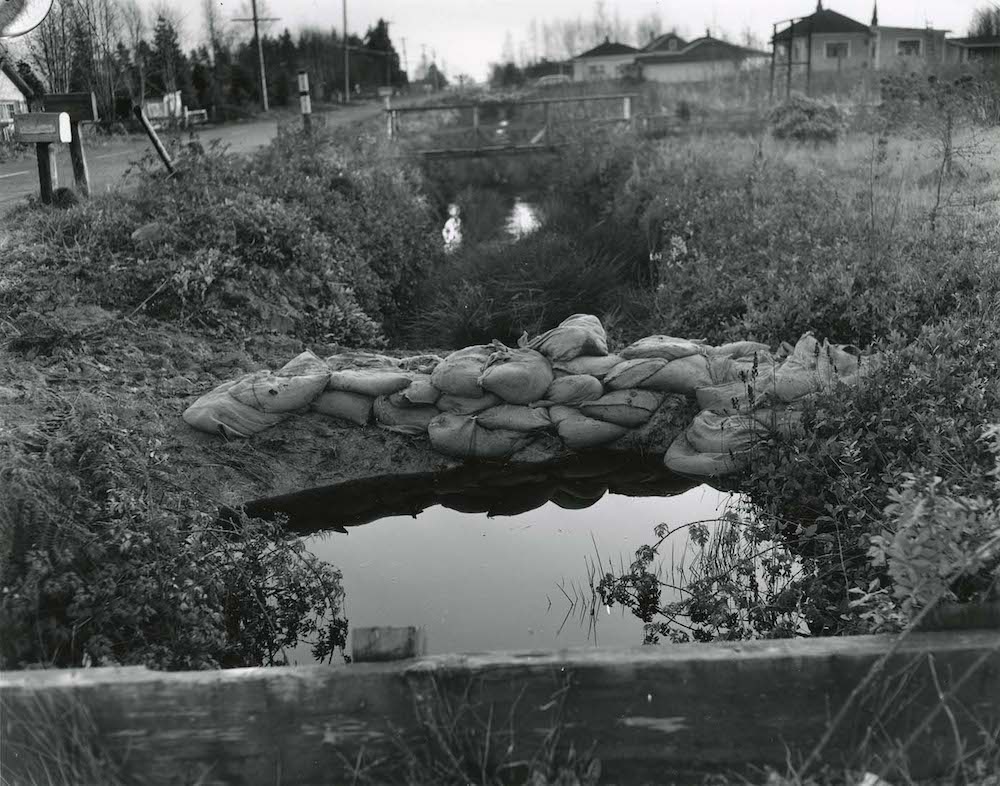 A black-and-white photo of flooded ditches by the side of the road.