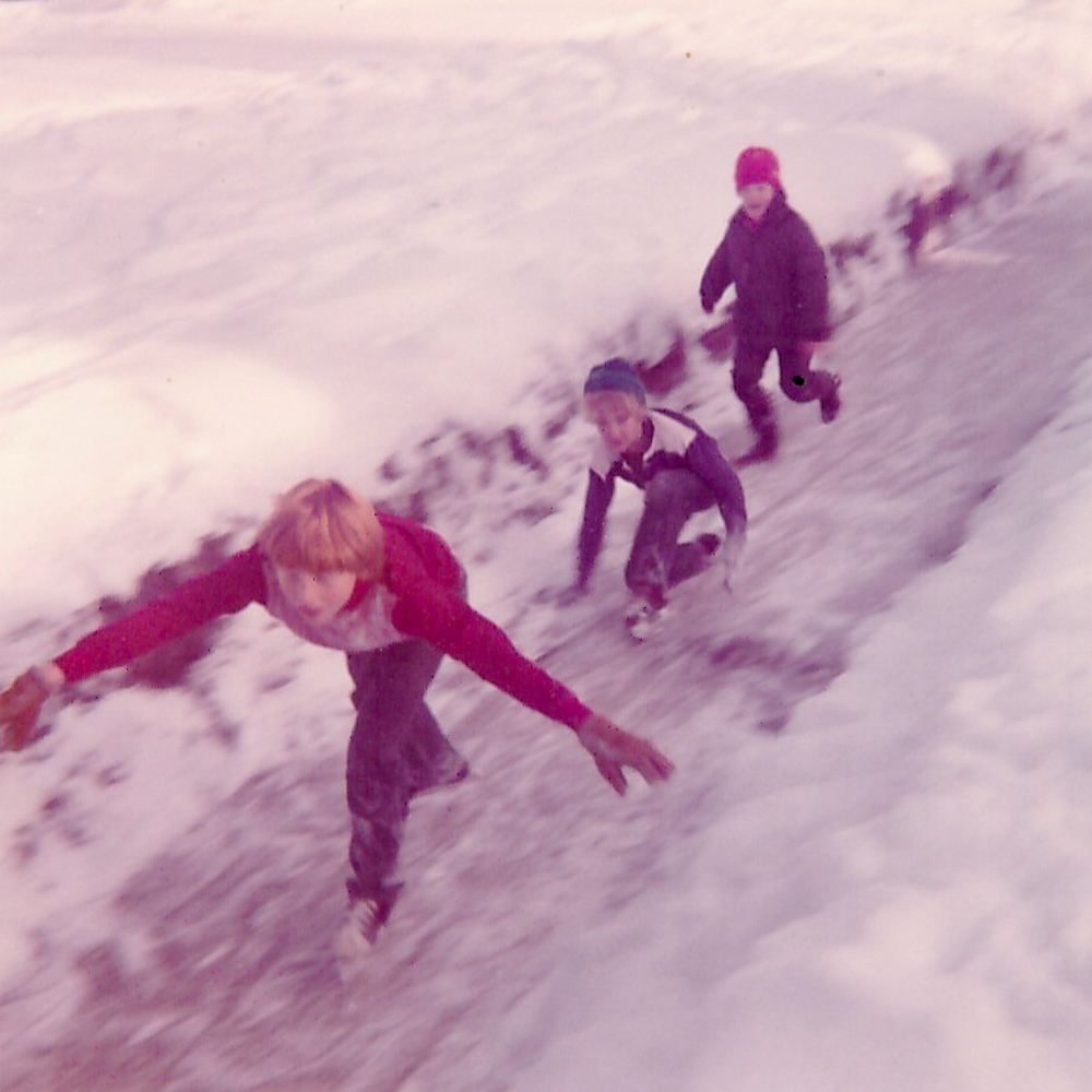 An old photo of three children in winter wear skating speedily down a frozen path.