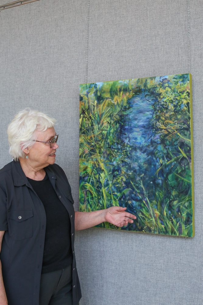 A woman with white hair stands beside a vertical painting on a canvas of a ditch with blue water and grassy sides.