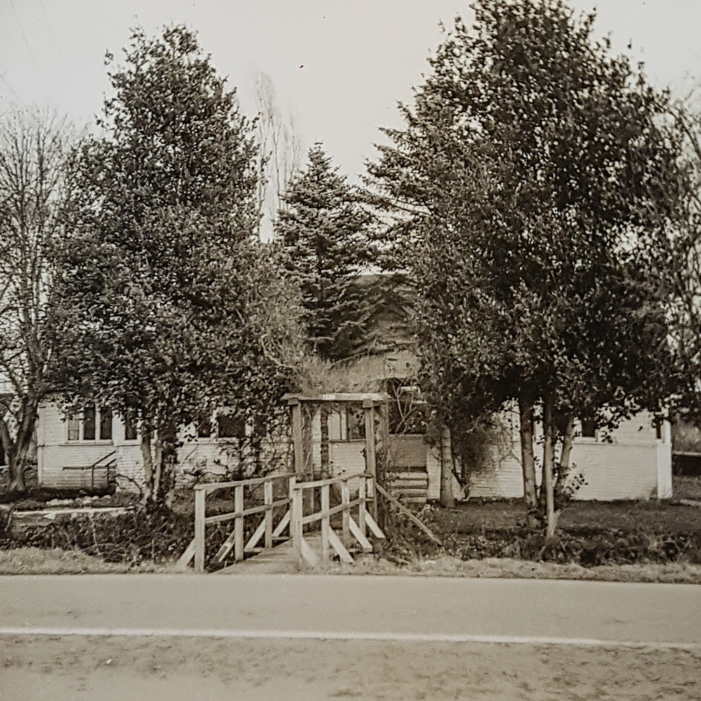 An old sepia photo of a wooden house with a wooden bridge over a ditch.