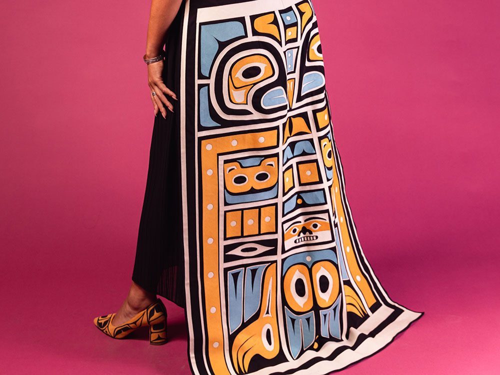 A black dress with brown fur trim that features a white, yellow, blue and black Northwest Coast train and cuffs and eagle artwork is worn against a fuschia background.