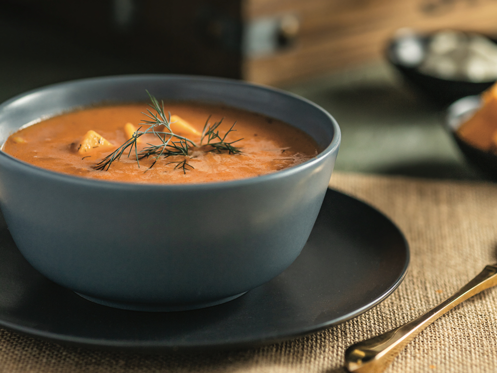 A bowl of borscht sits on a placemat on a table, with vegetables in the background.