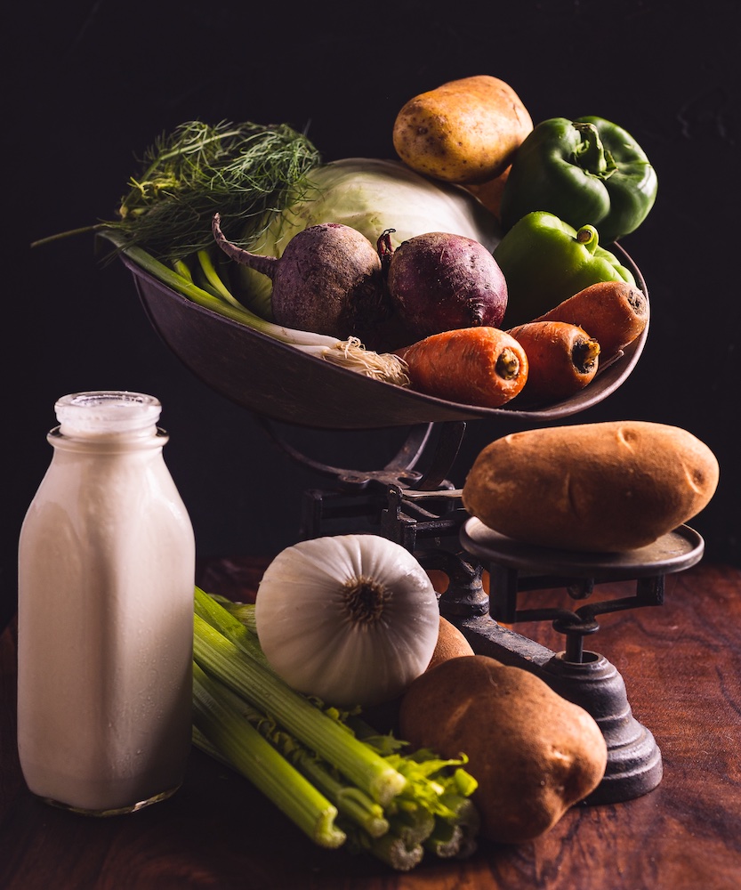 Ingredients to make electric pressure cooker Doukhobor borscht on a table. They include a glass bottle of heavy cream, garlic, dill, potatoes, beets, carrots, celery, and green pepper.