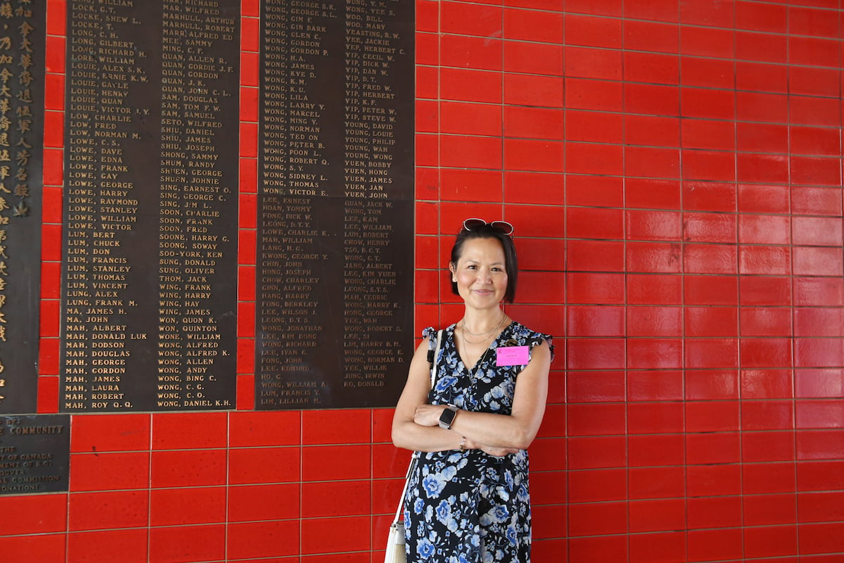 一名華裔婦女站在一麵紅瓷磚牆前，牆上掛著一塊刻有青銅名字的牌匾。