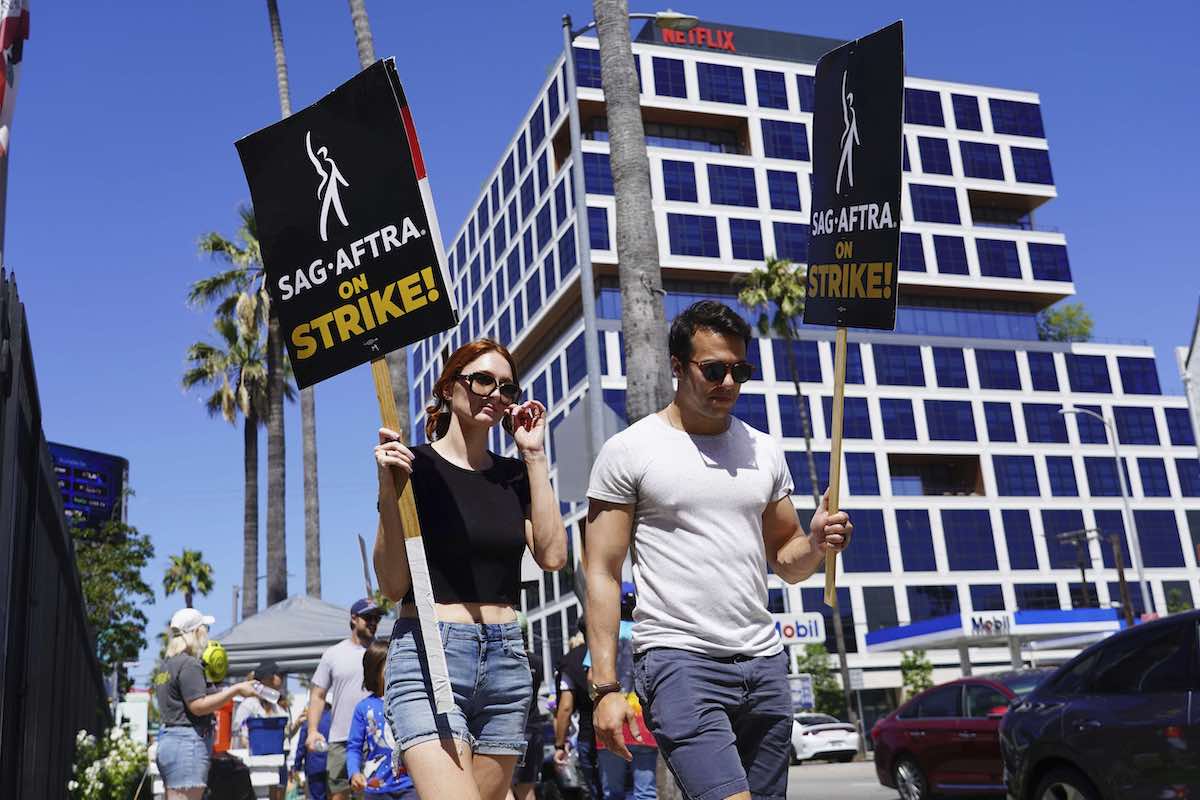 On the left, a woman with red hair, sunglasses, a black T-shirt and jean shorts holds a black picket sign that reads “SAG-AFTRA on strike!” She is walking beside a dark-haired man in sunglasses and a white T-shirt and grey shorts holding a similar sign. They are walking on the sidewalk in front of a large office building with square blue windows and cube-shaped white borders on a bright sunny day.