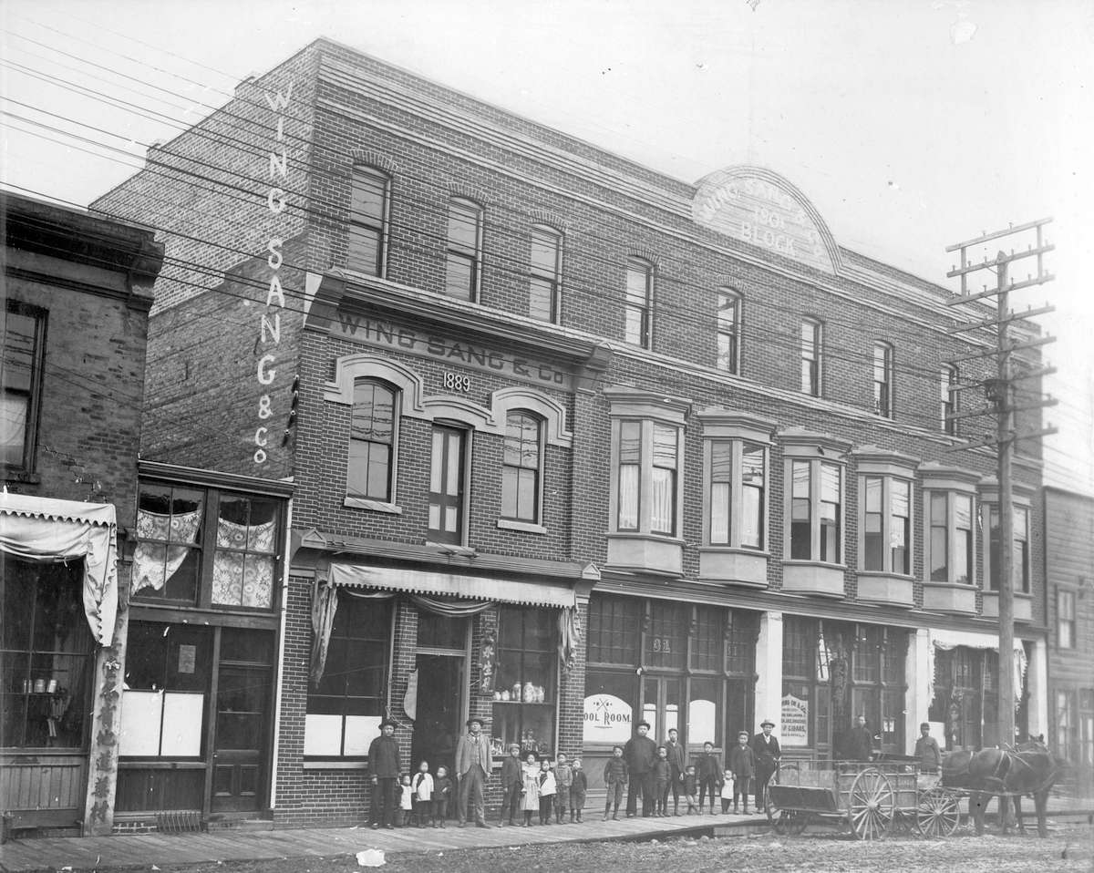 一張黑白照片，是一棟三層維多利亞式商業建築，前麵排著大約十幾個人，推著一輛馬車。