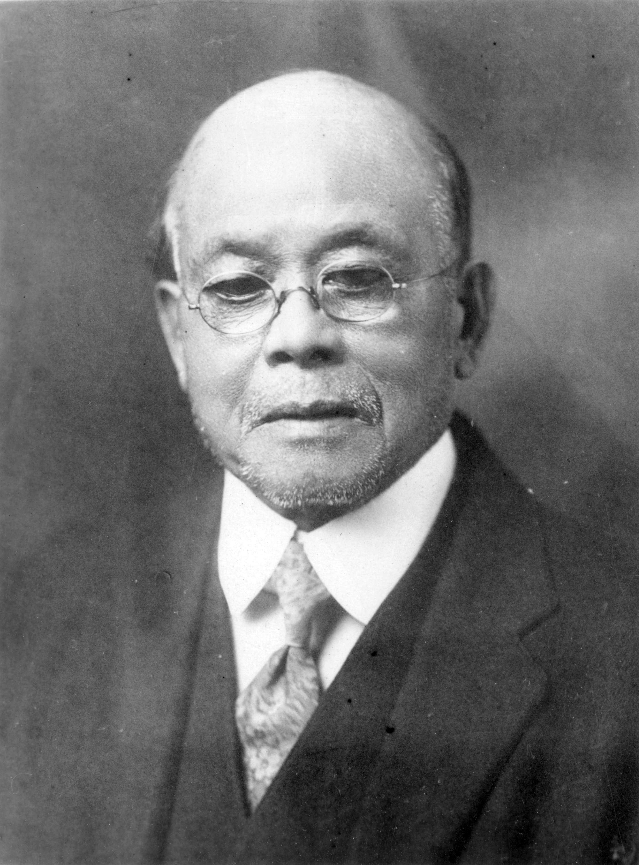 一位華裔老人的黑白肖像。 他戴著眼鏡，穿著西服。