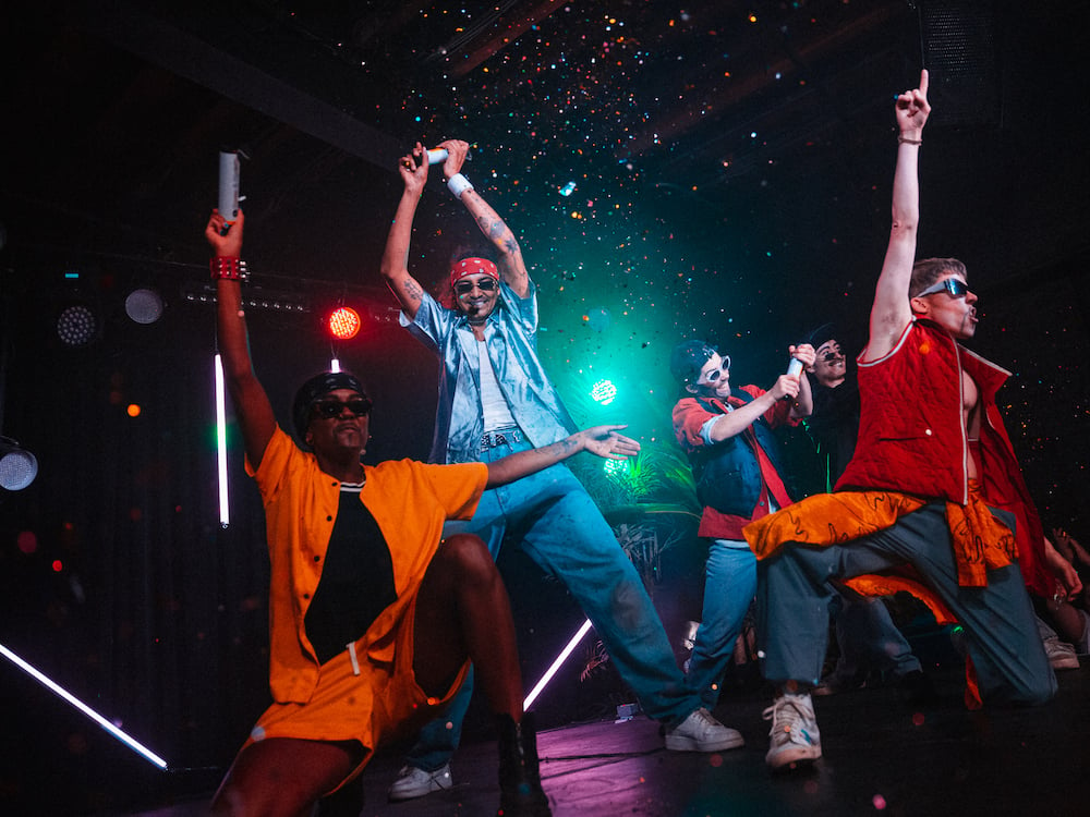 A group of smiling performers, dressed as a boy band, dance in the glow of a turquoise stage light as confetti fills the air.