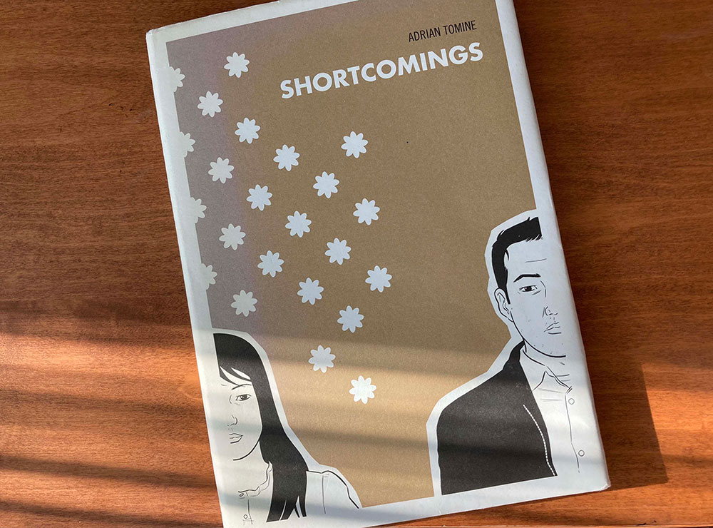 The book Shortcomings sits on a brown wooden tabletop. The cover features a black and white illustration of an Asian woman on the left and an Asian man on the right against a background that recalls brown kraft paper.